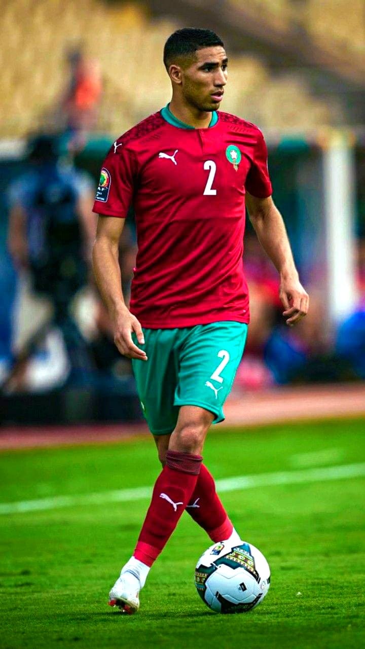A soccer player in a red and green Morocco jersey with number 2 dribbles a ball on the field during a match, as fans watching via IPTV Netherland follow every exciting moment.